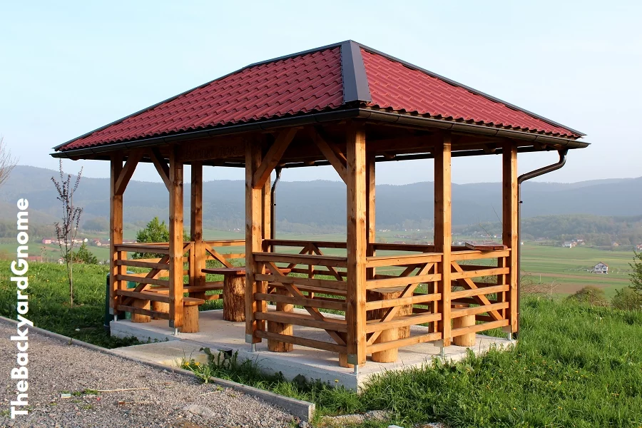 Newly built wooden gazebo structure with new roof mounted on concrete foundation with wooden table and chairs overlooking family houses and fields in distance with clear blue sky in background